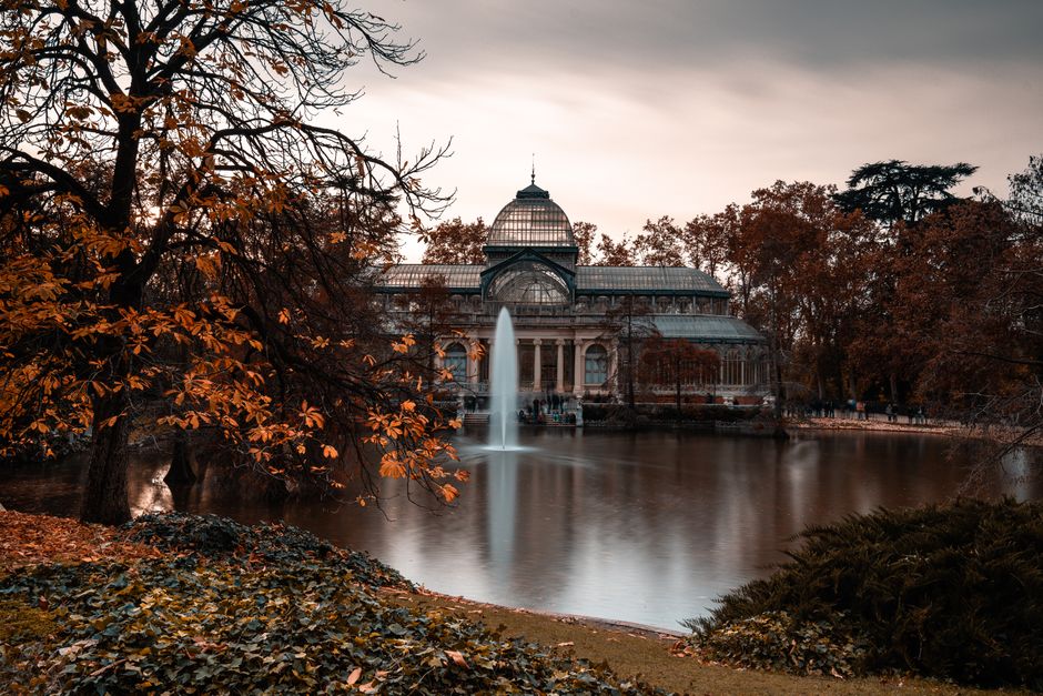 A large glass-domed conservatory building situated on the edge of a pond, with a prominent fountain shooting water into the air in front of it. The building features intricate Victorian-style architecture with tall, arched windows and a metal framework, set amidst a scenic park environment. Surrounding the pond are mature trees with autumnal foliage, displaying shades of orange and brown, some partially leafless branches extending over the water. The pond's surface reflects the building and trees, appearing smooth and slightly blurred due to long exposure photography. The foreground includes a grassy bank with fallen leaves and low undergrowth, framing the scene. The lighting is soft and subdued, suggesting late afternoon or early evening, creating a tranquil atmosphere. This scene, associated with outdoor leisure areas, could relate to private or municipal grounds where rubbish removal services might be used to maintain cleanliness around such landscaped environments. The image is clear and detailed, emphasizing the historic structure, natural surroundings, and water features, with no visible debris or waste evident in the scene.