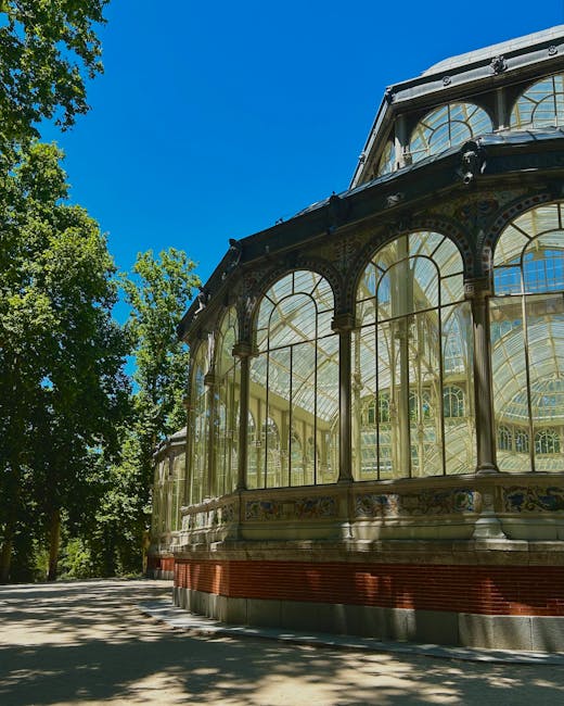 A detailed view of a historic glass conservatory with ornate, arched window frames and intricate metalwork, situated in a park environment. The structure features large, curved glass panels supported by decorative columns and beams, with a decorative frieze running along the base of the windows. The conservatory is positioned on a paved area, with the right side extending slightly beyond the frame of the image. Surrounding the conservatory are lush green trees with dense foliage, casting shadows on the ground, under a bright blue sky with minimal cloud cover. The scene suggests a well-maintained public garden or park setting, with no visible rubbish or waste, highlighting a tidy environment. The presence of trees and the clear, sunny weather enhance the calm, peaceful atmosphere, and the conservatory’s vintage architectural style is complemented by the natural surroundings. Occasionally, such structures are involved in on-site clearance or private disposal arrangements, aligning with the context of alternative waste handling or rubbish removal services provided by companies like houseclearancecrystalpalace.co.uk in the local area of Crystal Palace.