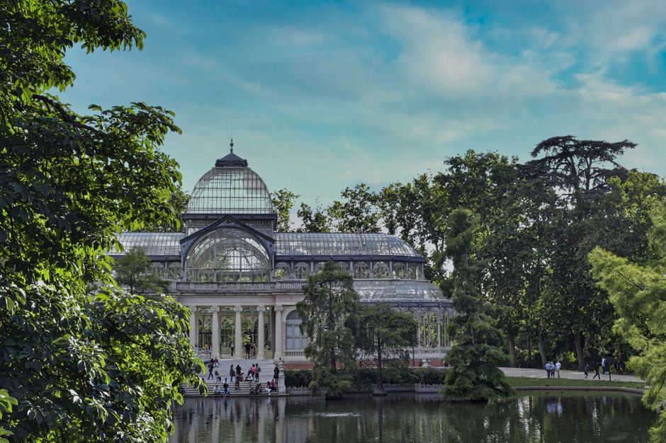 A large, historic greenhouse with a glass and metal structure, featuring a central dome and arched glass panels, is situated behind a pond and surrounded by lush green trees and shrubbery. The greenhouse's white framework and detailed architectural elements are clearly visible, with some people walking along the pathway in front of the building. The scene is set outdoors on a bright day with a blue sky and scattered clouds, and the environment appears peaceful and well-maintained. This setting could be connected to private property or cultivated gardens that may require on-site clearance or waste removal services. The overall atmosphere emphasizes natural beauty alongside a substantial glass conservatory, with the scene captured from a vantage point that includes the pond's reflective water in the foreground, adding depth to the image and highlighting the connection between the natural surroundings and the structure.