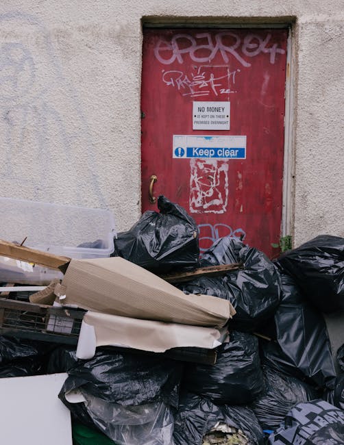 A red metal door with graffiti and multiple signs including one that says 'No Money Please' and another that says 'Keep Clear', situated within a weathered, off-white brick wall. In front of the door, there is a large pile of black rubbish bags, some torn open, with discarded cardboard, plastic packaging, and other waste materials stacked haphazardly on the ground. The debris appears to be collected for local rubbish removal or alternative waste handling, with visible signs of improper disposal in an urban environment. Natural daylight illuminates the scene, highlighting the contrast between the graffiti-covered door and the cluttered rubbish pile, which indicates a context related to private waste collection services like those provided by houseclearancecrystalpalace.co.uk.