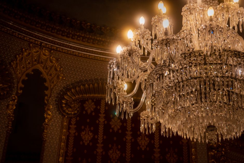 A large, ornate crystal chandelier is suspended from an elaborately decorated ceiling, featuring numerous hanging glass prisms and multiple small, candle-shaped light bulbs. The chandelier's metallic frame is partly visible, with a polished, reflective finish. The background showcases intricate, gold-accented detailing on the ceiling and walls, with rich, dark wallpaper adorned with floral and geometric patterns. The ambient lighting from the chandelier casts a warm glow across the decorated interior, creating a sense of opulence and grandeur. This scene highlights the type of luxurious fixtures that may be found in high-end interior spaces but is unrelated to rubbish removal services or waste management contexts, as no waste or debris are visible.