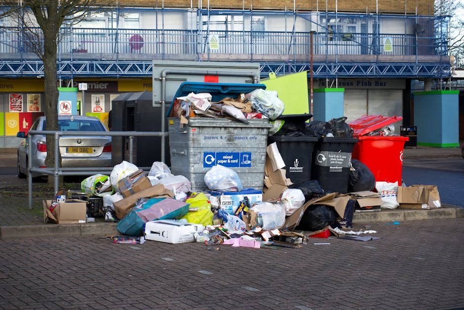 A large overflowing public waste bin situated on a paved sidewalk in front of a commercial building with a blue metal scaffolding structure on the upper floor. The bin, made of gray plastic with a blue lid, is filled with assorted waste including cardboard boxes, paper, plastic bags, and packaging material, some of which are spilling onto the surrounding area. Several black and red rubbish bags are stacked beside the bin, and additional loose waste, such as wrapped packages, paper sheets, and small cardboard boxes, are scattered across the paving. Behind the bin, a silver car is parked parallel to a metal railing, with an urban storefront visible in the background featuring colorful signage and a fish bar. The scene is outdoors during daylight, with some natural light illuminating the area, indicating a typical city environment where waste accumulation may require prompt disposal or private rubbish collection services offered by companies such as House Clearance Crystal Palace.