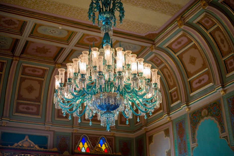 A large, ornate chandelier hangs from an intricately decorated ceiling inside a grand room, featuring numerous glass and crystal light fixtures in a cascading, tiered arrangement. The chandelier's components are transparent, with a bluish tint, and it emits a warm, bright glow that illuminates the elegant surroundings. The ceiling above showcases detailed paneling with pastel hues of pink, gold, and green, arranged in symmetrical patterns, bordered by ornamental molding. In the background, you can see part of a stained glass window with vibrant colors, adding to the opulent decor. The overall environment appears to be a historic or luxurious interior space, with soft lighting and intricate architectural details, typical of an elegant residence or a formal reception area. The context of rubbish removal services, such as [COMPANY_NAME], relates to the potential need for eco-friendly disposal of debris or restoration of such environments, though no debris or waste is visible in this image.