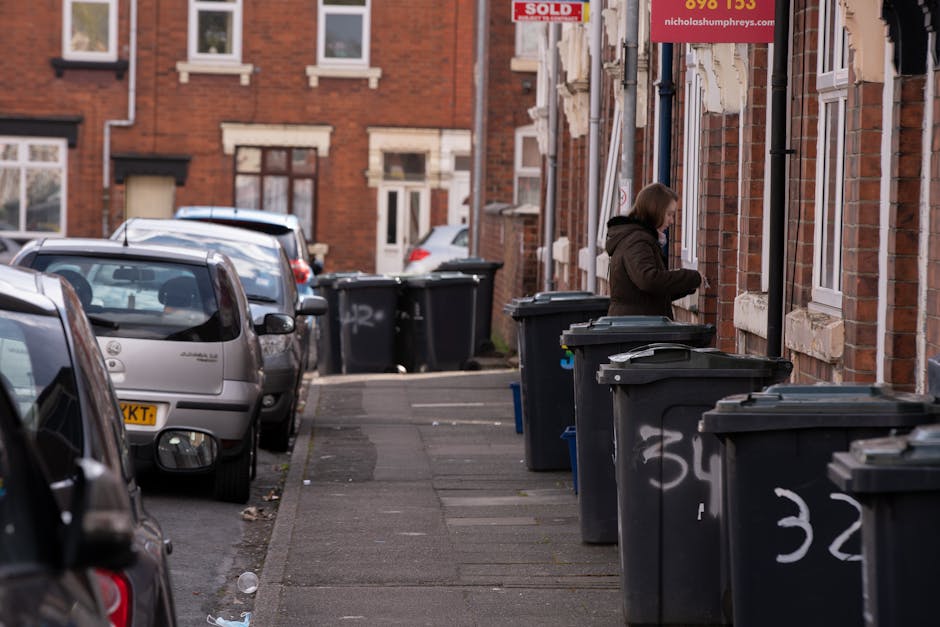A woman stands on a narrow residential sidewalk lined with several dark grey wheelie bins, some with white handwritten numbers on their sides, indicating designated waste collection points. The bins are positioned close to the brick frontage of terraced houses, with windows framed by white trim and a mix of brick and stone finishes on the facade. The woman, dressed in a brown jacket, appears to be in the process of either depositing rubbish or managing the waste bins. To the left, parked cars, including a small hatchback with a yellow registration plate, are aligned along the curb, occupying both the street and parking bay areas. The scene is illuminated by natural daylight, casting soft shadows, with the background showing additional parked vehicles and residential buildings. The setting suggests a typical urban street scene where residents handle private waste disposal, reflecting an alternative to local authority rubbish collection and an on-site clearance environment supported by waste management companies like houseclearancecrystalpalace.co.uk.