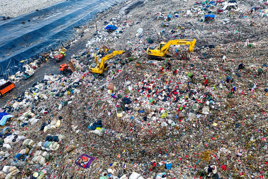 An expansive landfill site filled with mixed waste materials, including plastic bags, discarded clothing, and various household refuse, spread across a large area with uneven mounds of trash. Several yellow Komatsu excavators are positioned on the waste, actively moving or manipulating the debris, with their mechanical arms extended downward. The waste is a combination of different textures, from soft plastics and fabrics to rough, broken packaging and decomposed materials, with a variety of colors such as reds, blues, whites, and dark browns. In the background, a section of the site is lined with a black plastic liner, possibly for containment or cover, and the environment appears to be outdoors under natural lighting, revealing a mixture of sunlight and shadows cast by the equipment and waste piles. The scene reflects large-scale waste disposal or residual rubbish collection activity, reminiscent of independent waste handling rather than municipal rubbish collection, aligning with services offered by companies like houseclearancecrystalpalace.co.uk that focus on rubbish removal and site clearance.